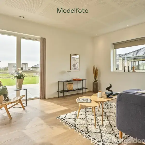 Stue med sofa og lænestol placeret på tæppe. Et sofabord står i midten. Store vinduer giver udsigt til have. Dekorationer inkluderer en lampe og planter. Tekst: "Modelfoto".