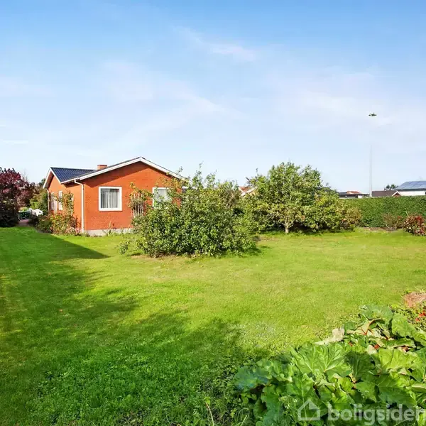 Et rødt hus står i en velplejet have med græsplæne og buske. Haven er omgivet af en hæk, og der er blå himmel i baggrunden.