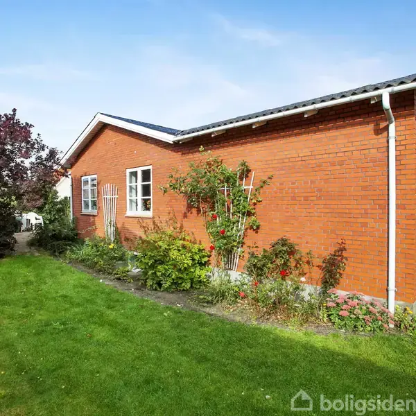 Rødt murstenshus står roligt omgivet af grønne buske og græsplæne, mens klatreplanter dekorerer muren. Skyfri himmel i baggrunden.