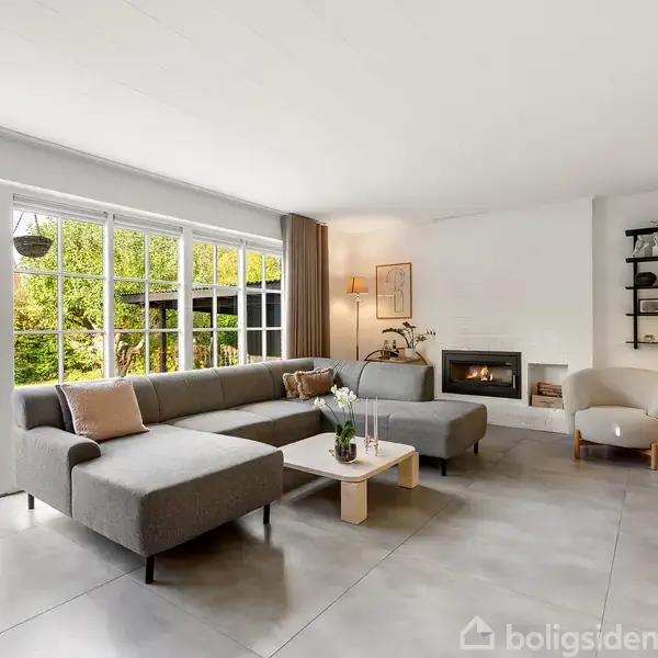 En L-formet grå sofa står i en lys stue med hvide vægge. Store vinduer viser en grøn have. En pejs er tændt ved siden af en lille lænestol.