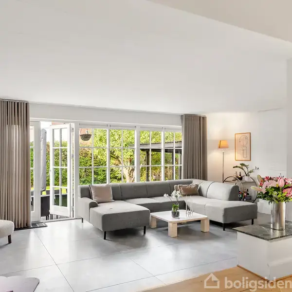 Stue med grå sofa i hjørne. Store vinduer lukker lys ind fra haven. Blomster og en lampe på bordet bidrager til en lys og indbydende atmosfære.
