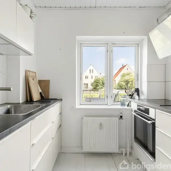 Et lyst køkken med hvide skabe og sorte bordplader. Der er en vask til venstre, ovn til højre, og et vindue i midten viser udsigt til huse.