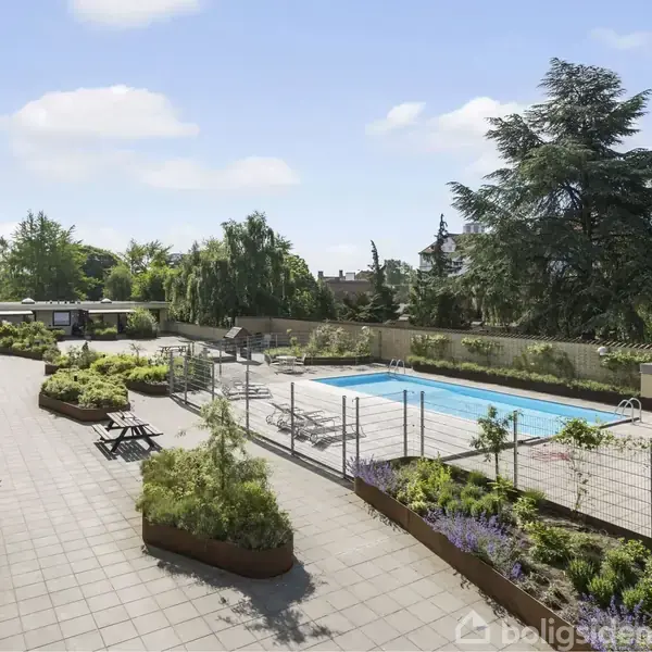 Udendørs swimmingpool står stille, omgivet af fliser og grønne planter i krukker; borde med bænke står langs stien, og høje træer omkranser området under en klar blå himmel.