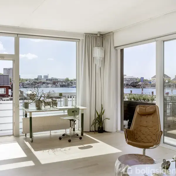 Et lyst værelse med store vinduer viser et skrivebord og stol mod en balkon. Udsigt over vandet og havnebygninger udenfor. Planter og illustrationer fuldender indretningen.