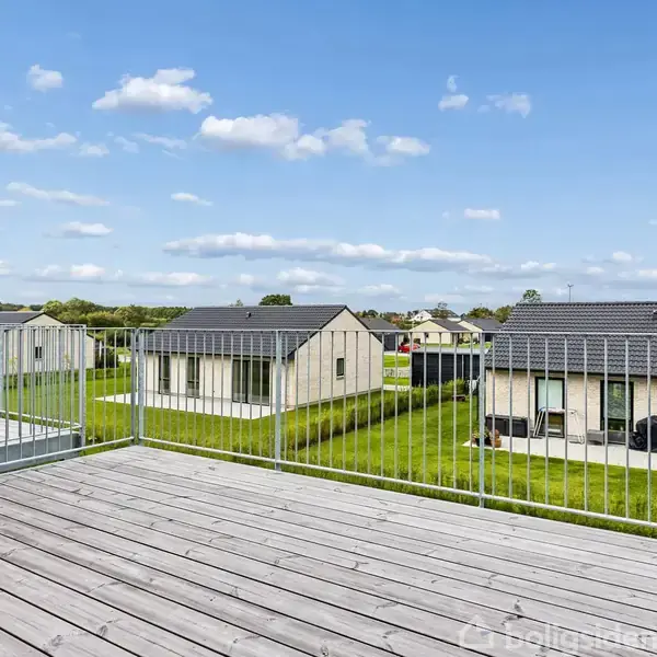 Træterrasse strækker sig foran huse med skrå tage. Græsplæner omkranser bygningerne, der ligger under en blå himmel med få skyer. Indhegning løber langs terrassekanten.