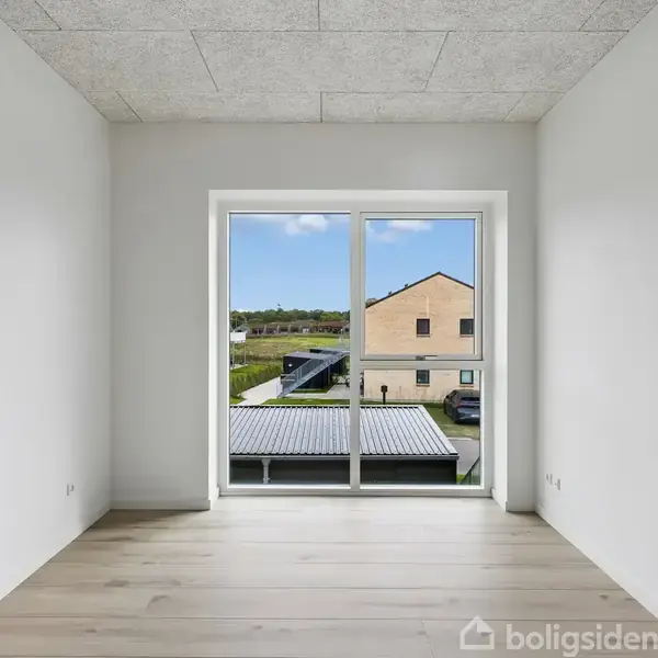 Et tomt rum med lyse trægulve og hvidmalede vægge, har en stor glasdør, der kigger ud over en gård med græs, huse og parkerede biler i det fjerne.