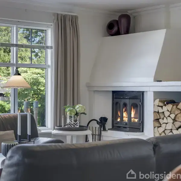 En pejs med brændende ild står i et hyggeligt opholdsrum, omgivet af polstrede møbler. Vinduet viser en grøn have udenfor, og en lampe ved siden af bordet giver belysning.