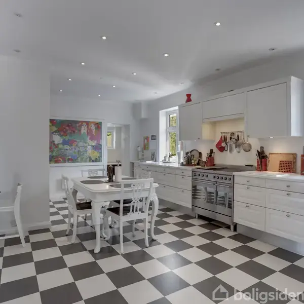 Et rummeligt køkken med et stort gaskomfur og hvide skabe strækker sig langs væggen. Et hvidt spisebord med stole står på et sort-hvidt ternet gulv i et lyst rum.