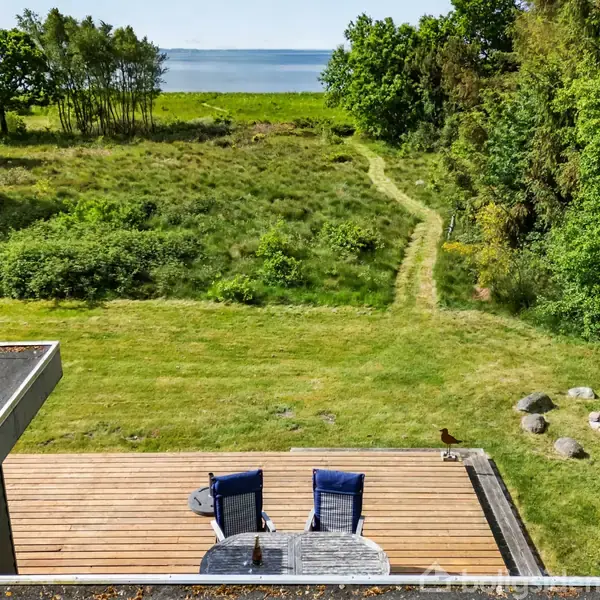 Terrasse med to stole og bord, orienteret mod græsplæne og sti, der fører til en skovkant og udsigt over vand i baggrunden.