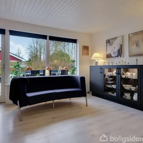 En stue med en sort sofa, der står foran store vinduer med udsigt til en terrasse og have. Væggene er lyse, og der er et sort skab med lamper og dekorationer.