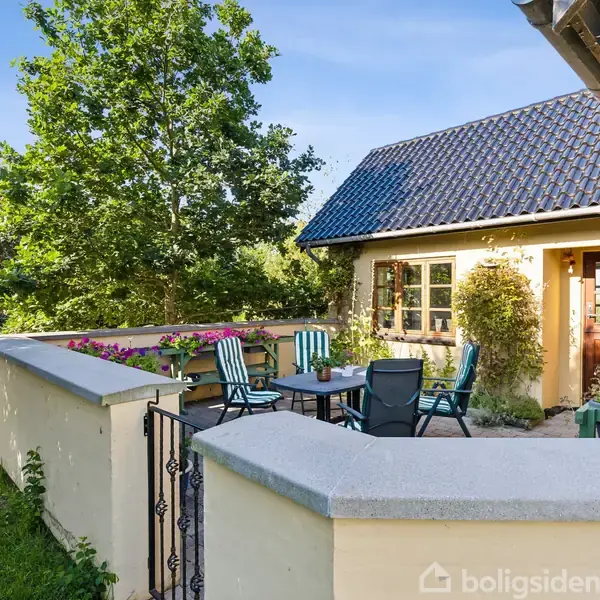 En terrasse er indrettet med havemøbler og blomsterkasser. Den støder op til et hus med blå tagsten, omgivet af træer og en græsplæne i en landlig have.