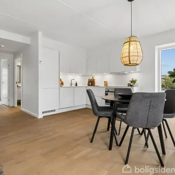 Spisebord med grå stole står i et moderne, lyst køkken. En stor lampe hænger over bordet. I baggrunden ses en sofa, planter og en dør til altanen.