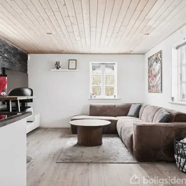 Brun sofa i en stue med et træloft, placeret ved to vinduer. Rundt sofabord står på et tæppe. Køkkenbord med stole til venstre. Kunst på væggene.