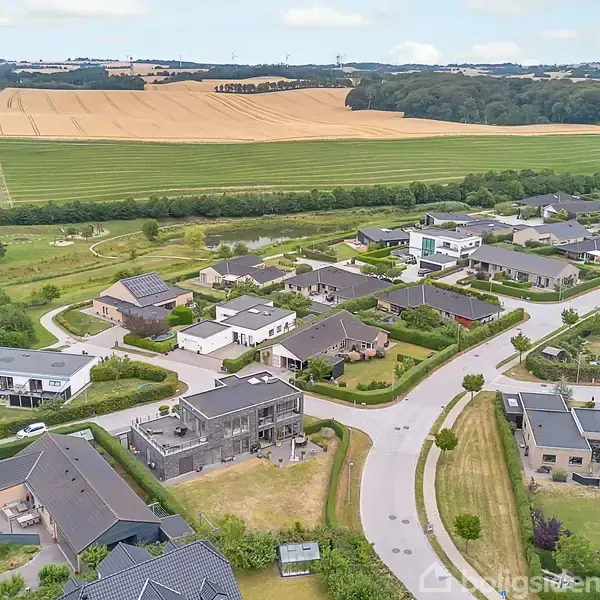 Bebyggelse set fra luften, viser moderne huse og snoede veje omgivet af grønne områder og marker, der strækker sig ud i det fjerne, under en delvis skyet himmel.