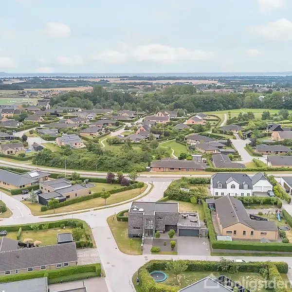 Et boligområde med rækkehuse og små veje omgivet af grønne arealer. Området ligger i et åbent landskab med marker og træer i baggrunden under en lyseblå himmel.