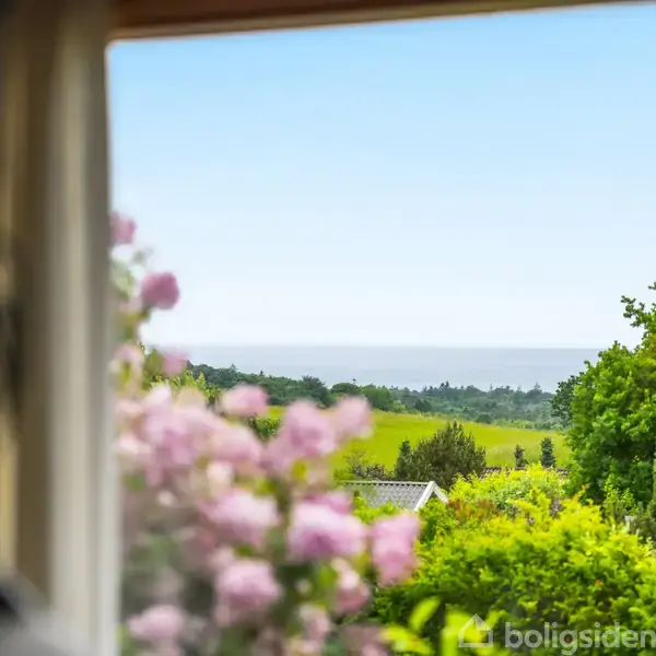 Blomster sidder i en vindueskarm, med udsigt over en frodig have og landskab, hvor havet ses i horisonten under en klar blå himmel.