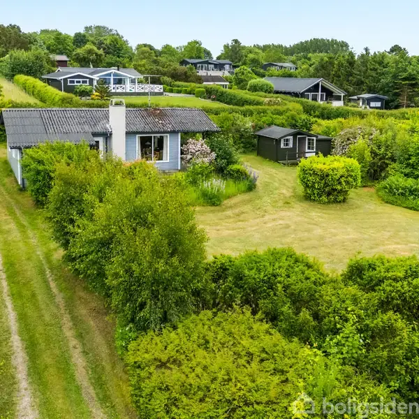 Fritidshus med sort tag står stille omgivet af grønne buske og græsplæne. En grussti fører op til huset, og flere huse er synlige i baggrunden.