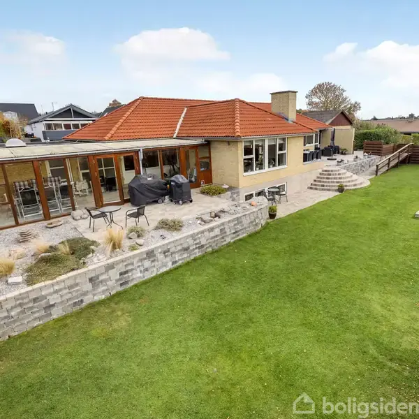 En gul murstensvilla med rødt tegltag står i en velholdt have. Der er en stor terrasse med havemøbler og grill, omgivet af grøn græsplæne og stengærde.