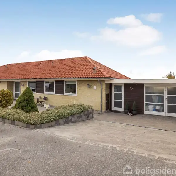 Et enkelt enfamiliehus med rød tegltag ses fra fronten. Huset har gule mursten, en lille forhave og en carport med plads til affaldsbeholdere. Himmel med enkelte skyer.