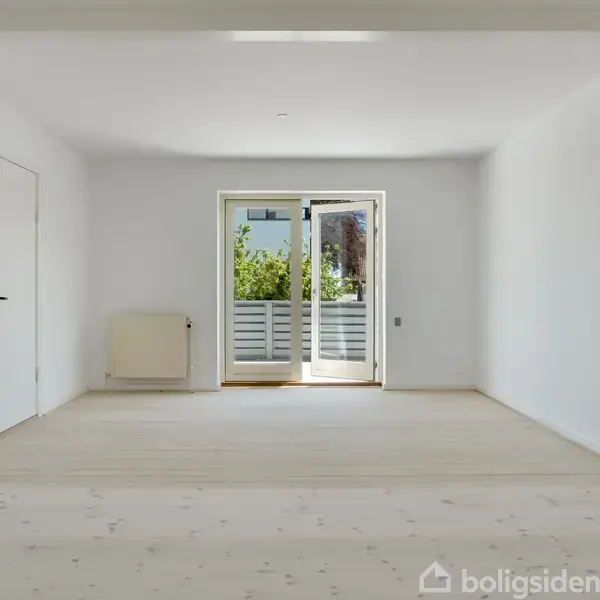 Et lyst og tomt rum fører ud til en lille balkon gennem en dobbeltdør. Væggene og loftet er hvide, og der er et varmeapparat ved venstre væg.