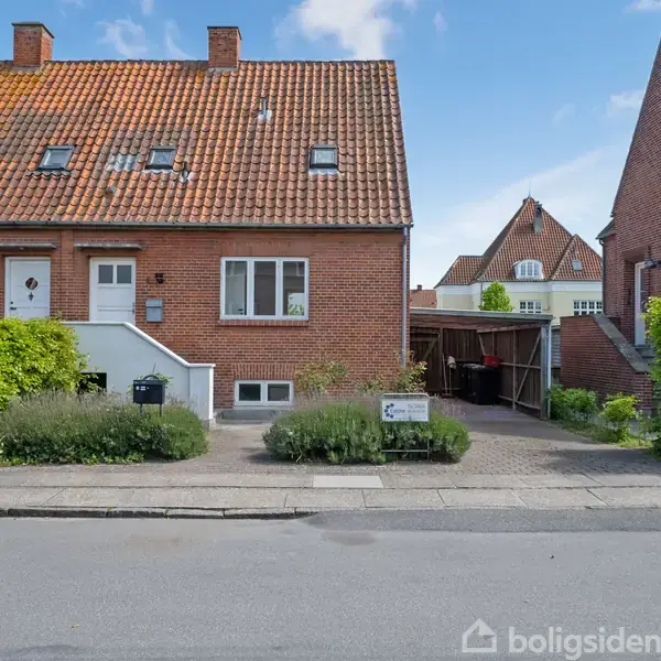 Tofamiliehus i røde mursten med rødt tegltag ligger i et kvarter med lignende bygninger og haver. Skilt ved indkørsel viser "Til salg".
