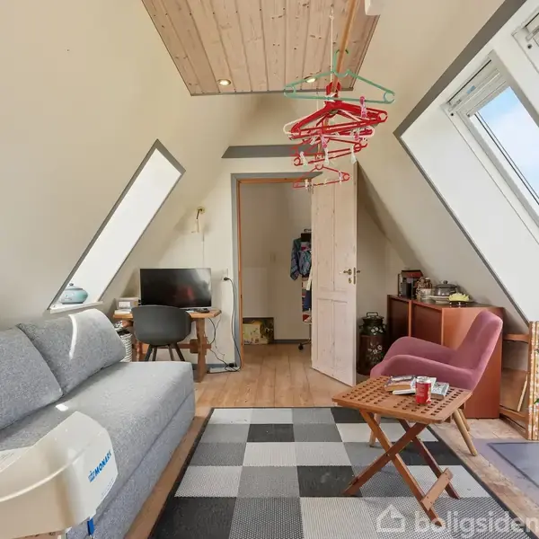 Et spisebord med seks stole er centralt placeret under en loftslampe. Rummet har træloft, hvide vægge, hylder med dekorationer og store vinduer, der giver udsigt til en grøn have.