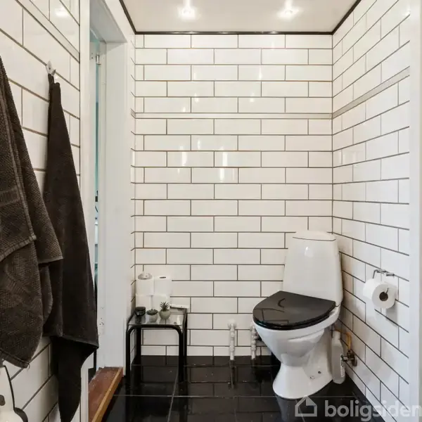 Toilet står i et rum med hvide fliser på væggene og sort gulv. Ved siden af hænger to mørke håndklæder på kroge. Rummet har en lys og ren atmosfære.
