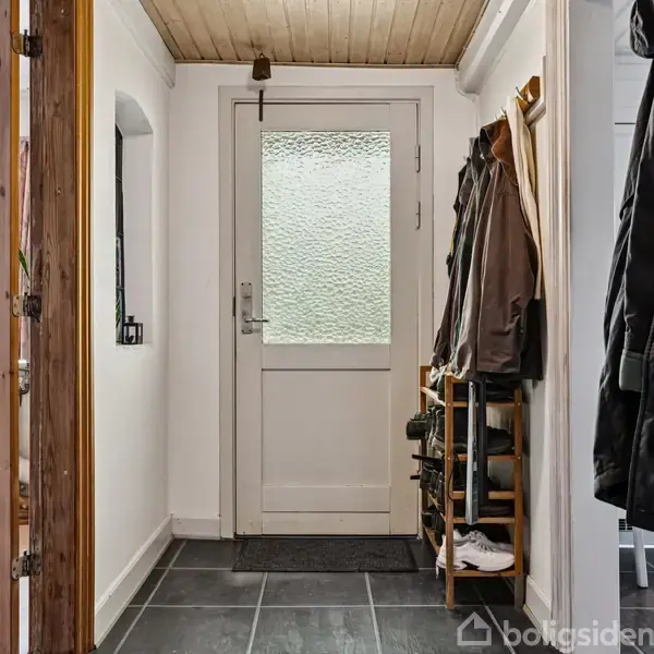 Dør med matteret glas fører ind til en entré med mørke fliser. Til højre står en knagerække fyldt med jakker, og et skoskab til opbevaring ses ved siden af.