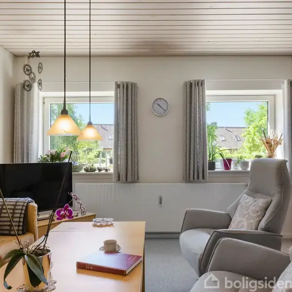 Stue med sofa og to lænestole omkring et bord. En tv-skærm står ved siden af planter i vinduer. To hængelamper lyser rummet, der har en neutral, lys indretning. En ur hænger på væggen.