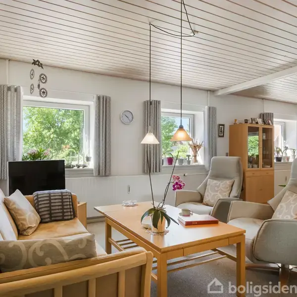 En stue med lyse træmøbler; sofa og lænestole omkring et sofabord. Rummet er lyst med store vinduer, udsigt til grønne træer, og dekoreret med lamper og planter.