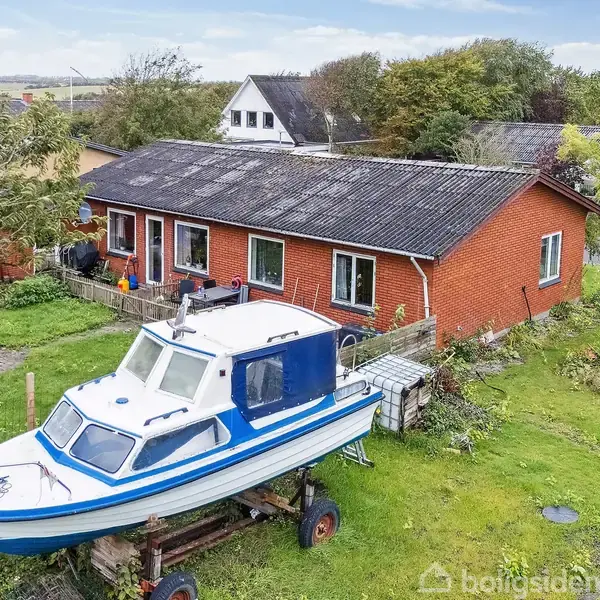 En båd står på en trailer foran et rødt murstenshus med flere vinduer. Haven er grøn og frodig med træer og buske, omgivet af et boligområde.