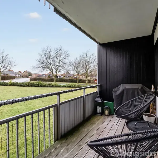 To stole står på en altan med trægulv, grønne plæner og nogle boliger i baggrunden. Der er et bord ved siden af stolerne og en gasgrill dækket af et mørkt betræk.