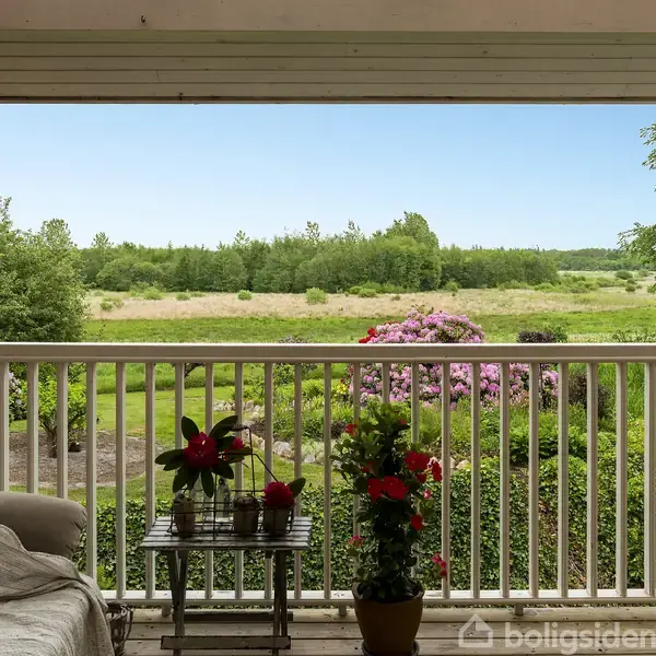 En sofa står på en overdækket terrasse med et bord og planter. Udsigt over grøn have med blomster og åbent landskab med træer i baggrunden.