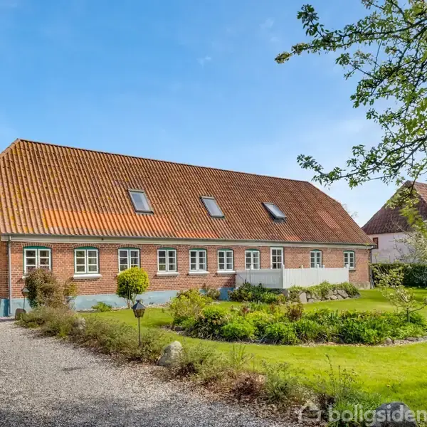 Et rødt murstenshus med skråt tegltag står i en frodig have omgivet af grussti og grønne træer under en klar blå himmel, der skaber en idyllisk scene.