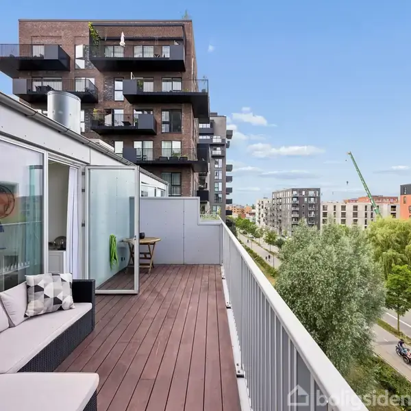 Altan med trædæk og beige sofa, beliggende ved moderne lejlighedsbygninger i en bymæssig bebyggelse. Udsigt over veje, træer og flere etagebyggerier under en klar blå himmel.