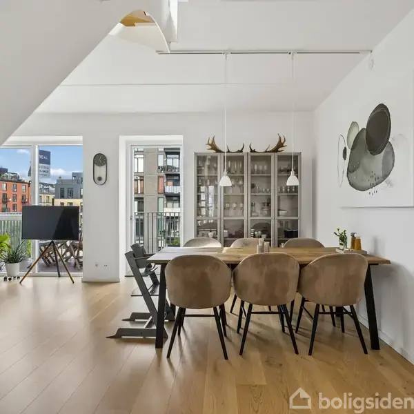 Spisebord med seks polstrede stole står i et moderne rum med trægulv. Et glasvitrine med dekorative genstande i baggrunden, omgivet af vinduer med udsigt til en urban skyscape.