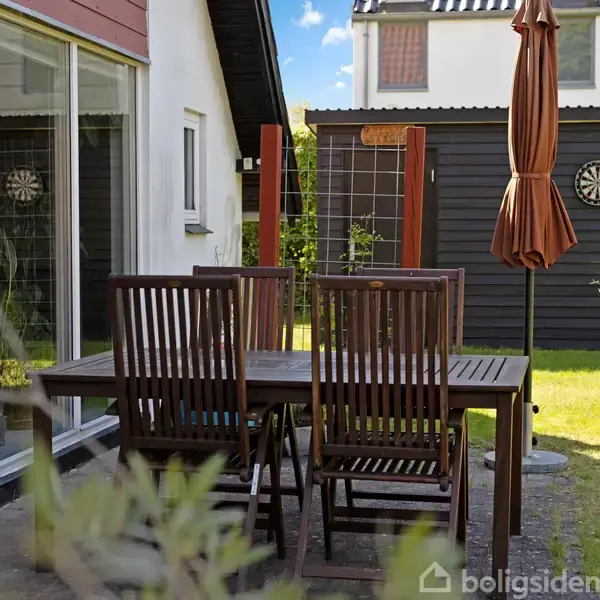 Træbord med fire stole står på en terrasse foran et hus. En lukket parasol ses ved siden af bordet. I baggrunden er en dartskive på et sort træhus.