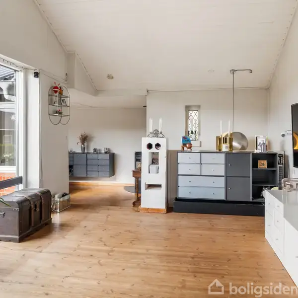 Stue med trægulv har et moderne interiør, bestående af sorte og hvide møbler, herunder en væghængt TV-enhed. Store vinduer til venstre giver udsigt til en terrasse med murstensvæg.