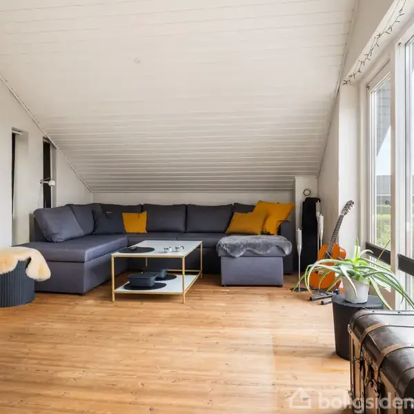 En stue har en stor, grå hjørnesofa med gule puder. Et sofabord står foran sofaen. Rummet er lyst med store vinduer og har træplankegulv. En plante og en guitar står ved vinduet.