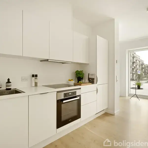 Et lyst køkken med hvide skabe, hvor en ovn og vask er integreret. Store åbne døre leder til en balkon med udsigt til en boligbygning.