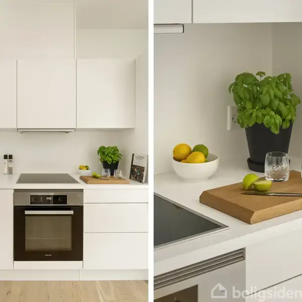 Hvidt køkken med indbyggede skabe og ovn; en skål med citroner, en grøn plante og en opskriftsbog står på bordet ved siden af glas og en skærebræt med limefrugt.