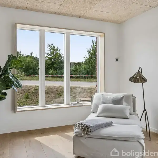 En hvid chaiselong står ved et stort vindue med udsigt til grønne træer. En gulvlampe står ved siden af, og der er en stor plante i en hvid vase til venstre.