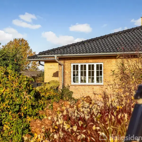 Et gult murstenshus med sorte teglsten, omgivet af frodig efterårsbevoksning, ses i sollys. Forgrund med forskelligartede buske og blå himmel i baggrunden.
