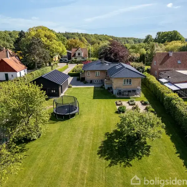 Hus med blått tag står i en stor, velholdt have med grønt græs. En trampolin er placeret på plænen ved siden af mindre træer og buske. Omkringliggende boliger og grønne områder ses i baggrunden.