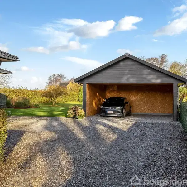 Carport med en sort bil parkeret, omgivet af en grusbelagt indkørsel og grøn hæk. I baggrunden ses et velplejet græsareal og husets murstensvæg.