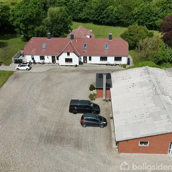 Et hvidt hus med rødt tegltag står mellem træer, omgivet af en stor grusplads. Flere biler er parkeret, og en rød murstensbygning med gråt tag ligger ved siden af.