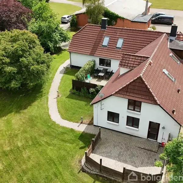 Hus med rød tag udført i tegl, omkranset af en grøn have. Flisebelagt terrasse foran huset. Ved siden af huset ses en grusvej.
