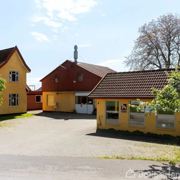 To bygninger med gule facader og røde tagsten står i en gård, omgivet af grønne træer og en klar blå himmel. Asfalteret vej leder til gården.