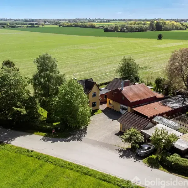 En gul bygning omgivet af grønne træer står stille ved en landevej, indrammet af frodige marker, der strækker sig mod horisonten under en klar blå himmel.
