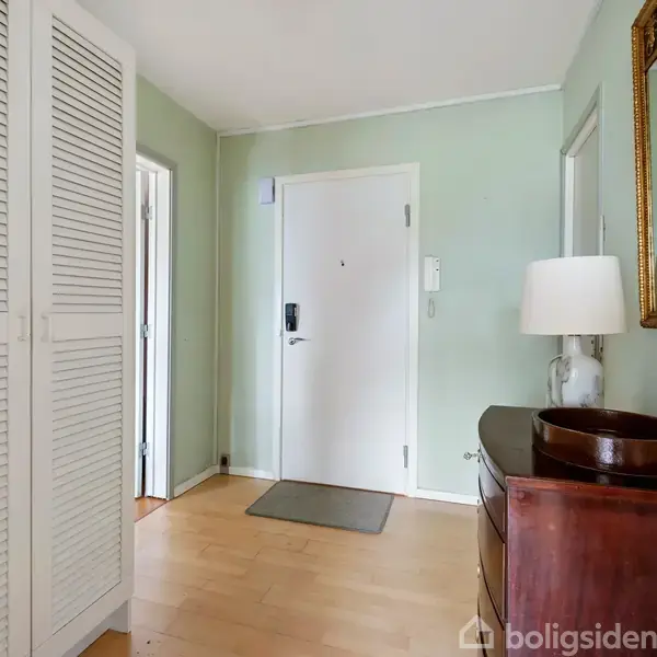 Spejlreflekterende indgangsparti med hvid dør, beige vægge og lys trægulv. Til venstre står et skab. En kommode med lampe og skål står til højre under spejlet.